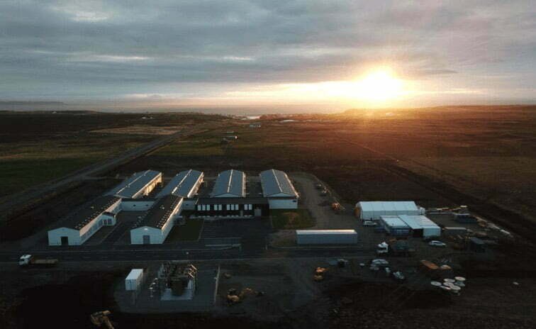 Ilustracja przedstawia kopalnię kryptowalut w miejscowości  Blonduos. Widoczne są podłużne hale przemysłowe, dużo samochodów oraz dróg dojazdowych.