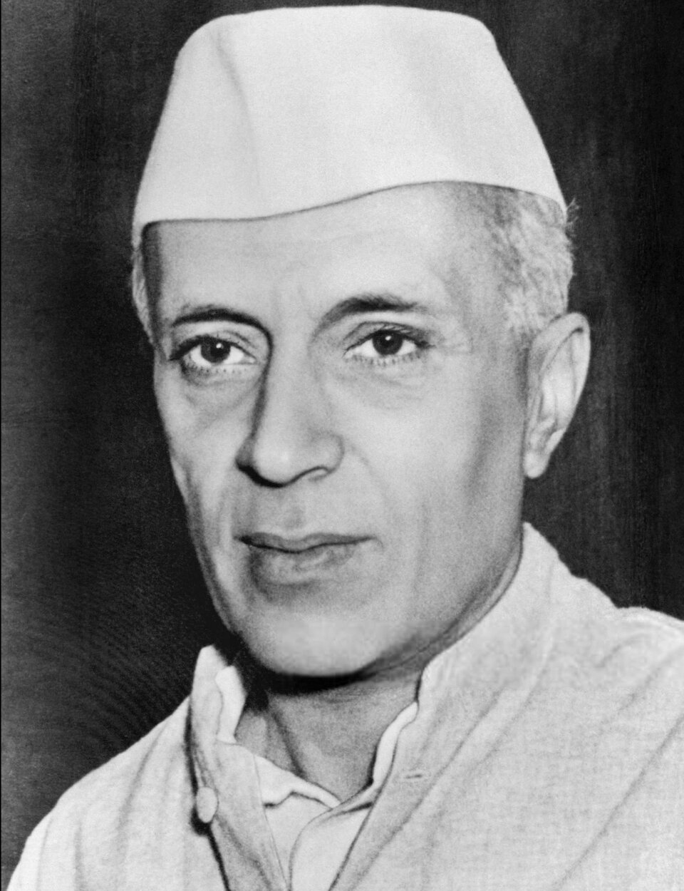 Na czarno‑białym zdjęciu znajduje się Jawaharlal Nehru. Ma on krótkie siwiejące włosy i jest ubrany w białą koszulę i czapkę. 