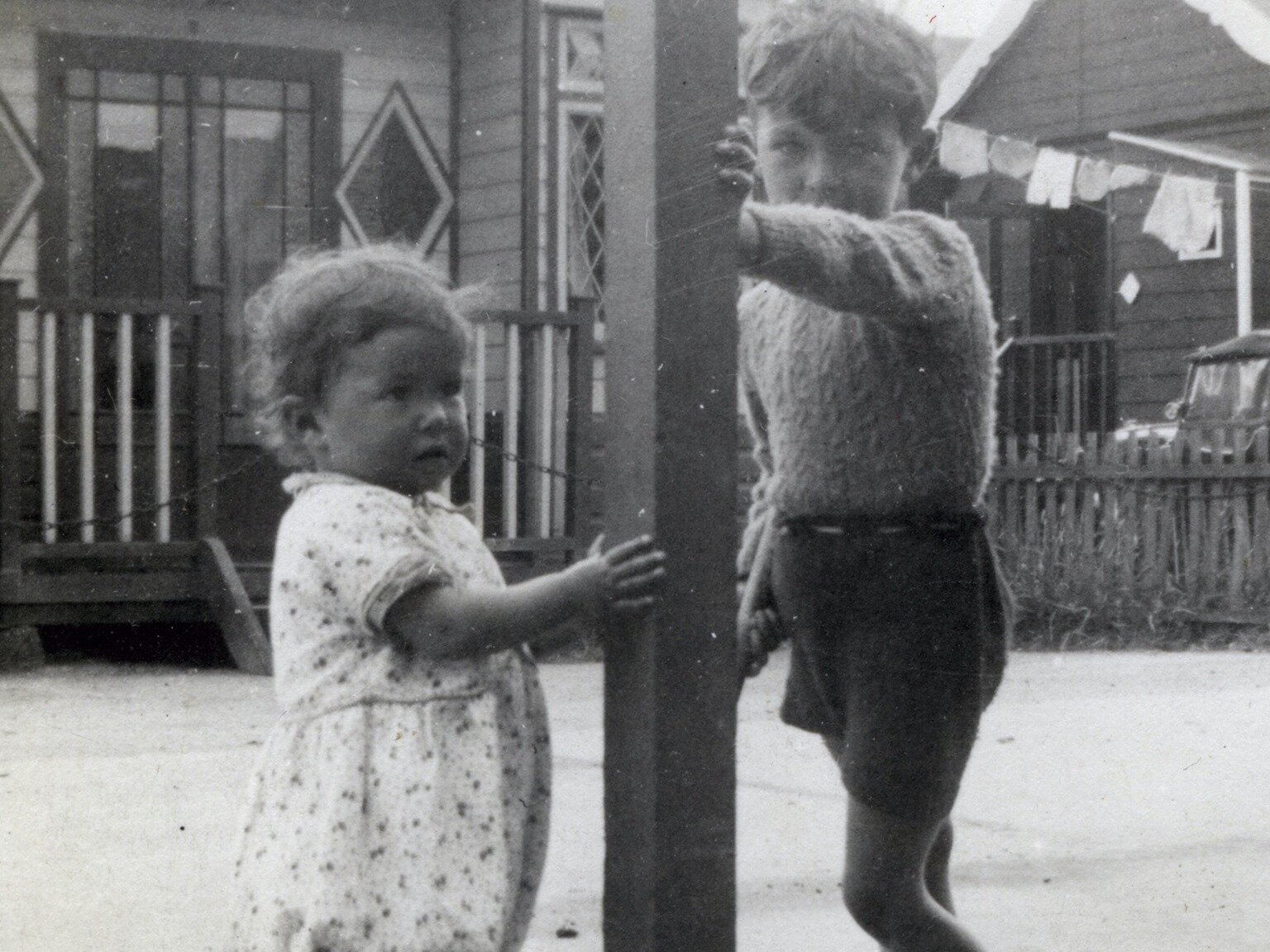 Czarno‑białe zdjęcie przedstawia dwoje dzieci opierających się rękoma o słup. Po lewej stronie widoczna jest dziewczynka w wieku około 2 lat, ubrana w jasną sukienkę. Po prawej stronie widoczny jest chłopiec w wieku przedszkolnym, ubrany w ciemne, krótkie spodnie, w które ma włożony sweter. W tle widoczne drewniane domy z otwartymi werandami, drewniany płot, pranie suszące się na sznurze.