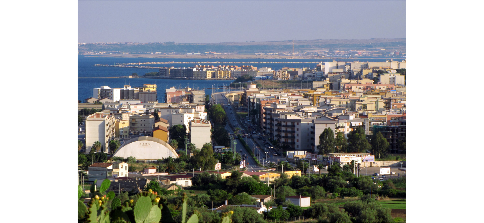 Zdjęcie przedstawia widok na panoramę sycylijskiego miasta Augusta. Zdjęcie wykonane jest z góry, widoczne są budynki, a w oddali morze.