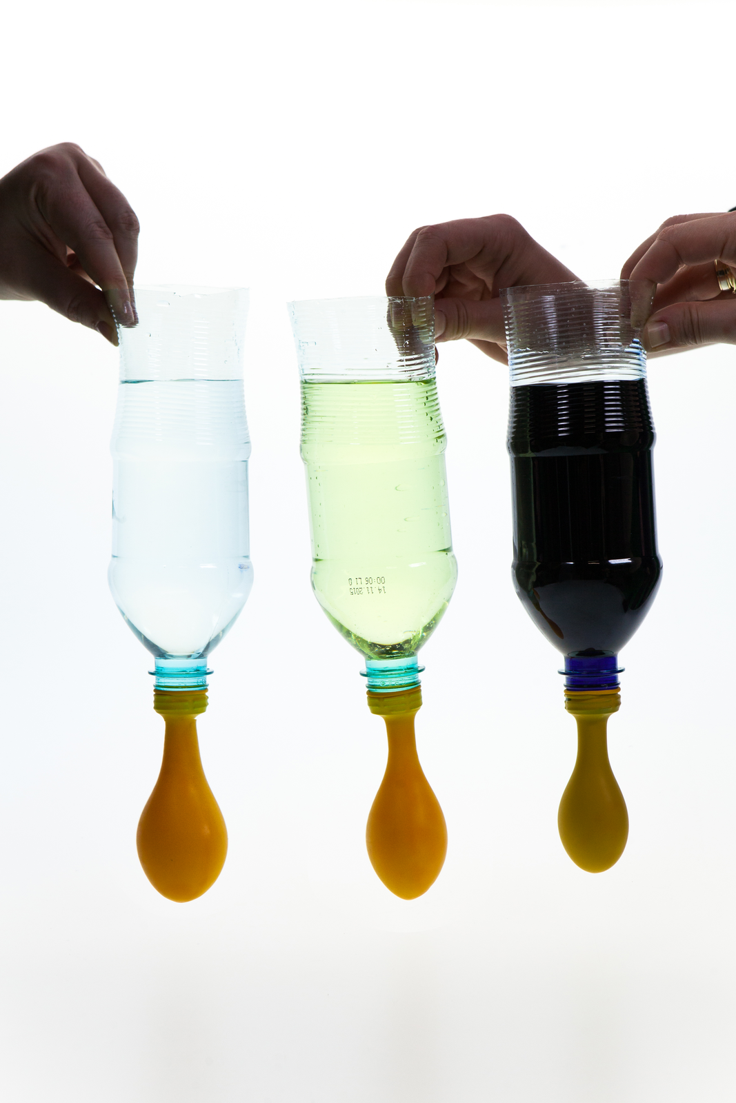 Zdjęcie przedstawia trzy dłonie trzymające butelki ze ściętymi dnami, od strony ściętego dna. Tło białe. W butelkach są różnokolorowe ciecze: w lewej przezroczysta i bezbarwna, w środkowej przezroczysta i lekko żółta, w prawej nieprzezroczysta i czarna. Na gwinty butelek naciągnięte są balony, wyglądające na lekko nadmuchane. Balon naciągnięty na lewą butelkę jest najbardziej nadęty, balon naciągnięty na prawą butelkę jest najmniej nadęty.