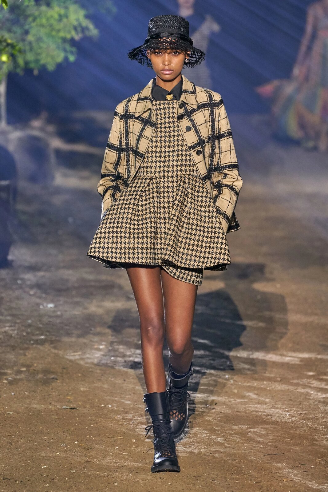 Fotografia przedstawia ciemnoskórą modelkę, która ma na sobie czarno‑białą rozkloszowaną sukienkę z wzorem w pepitkę spod której wystaje ciemniejszy koszulowy kołnierz, kraciastą kurtkę w kolorze beżowym z domieszką szarości, czarne botki do połowy łydki oraz czarny kapelusz przykrywający włosy. Sukienka odsłania szczupłe nogi kobiety. Jej wzrok skierowany jest do przodu, a dłonie ukryte w kieszeniach kurtki.