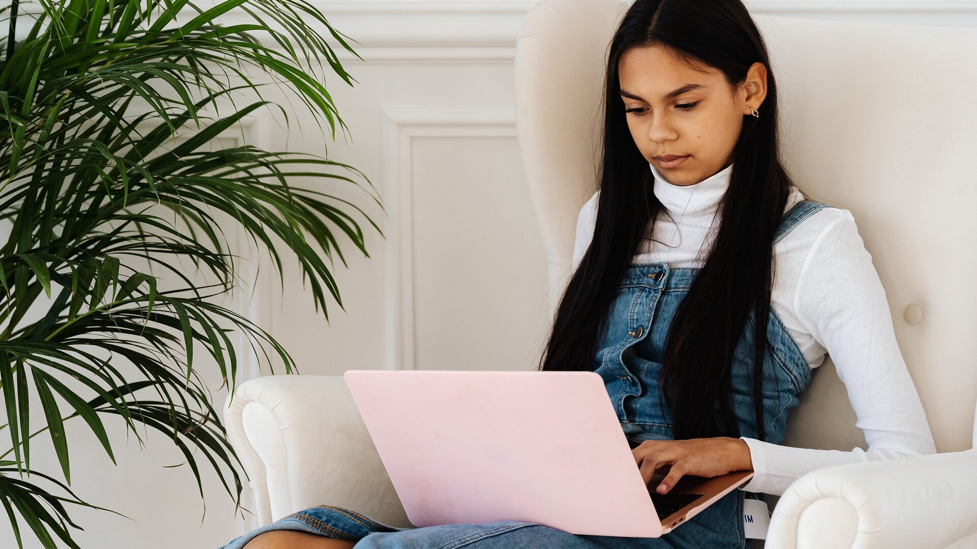 Zdjęcie przedstawia nastolatkę, która siedzi na dużym białym fotelu i wpatruje się w otwarty, różowy laptop. Dziewczyna ma długie, czarne włosy, jest ubrana w biały golf i dżinsowe ogrodniczki. Jej głowa jest lekko pochylona, a lewa ręka spoczywa na klawiaturze laptopa. Po lewej stronie widoczny jest duży zielony kwiat doniczkowy.
