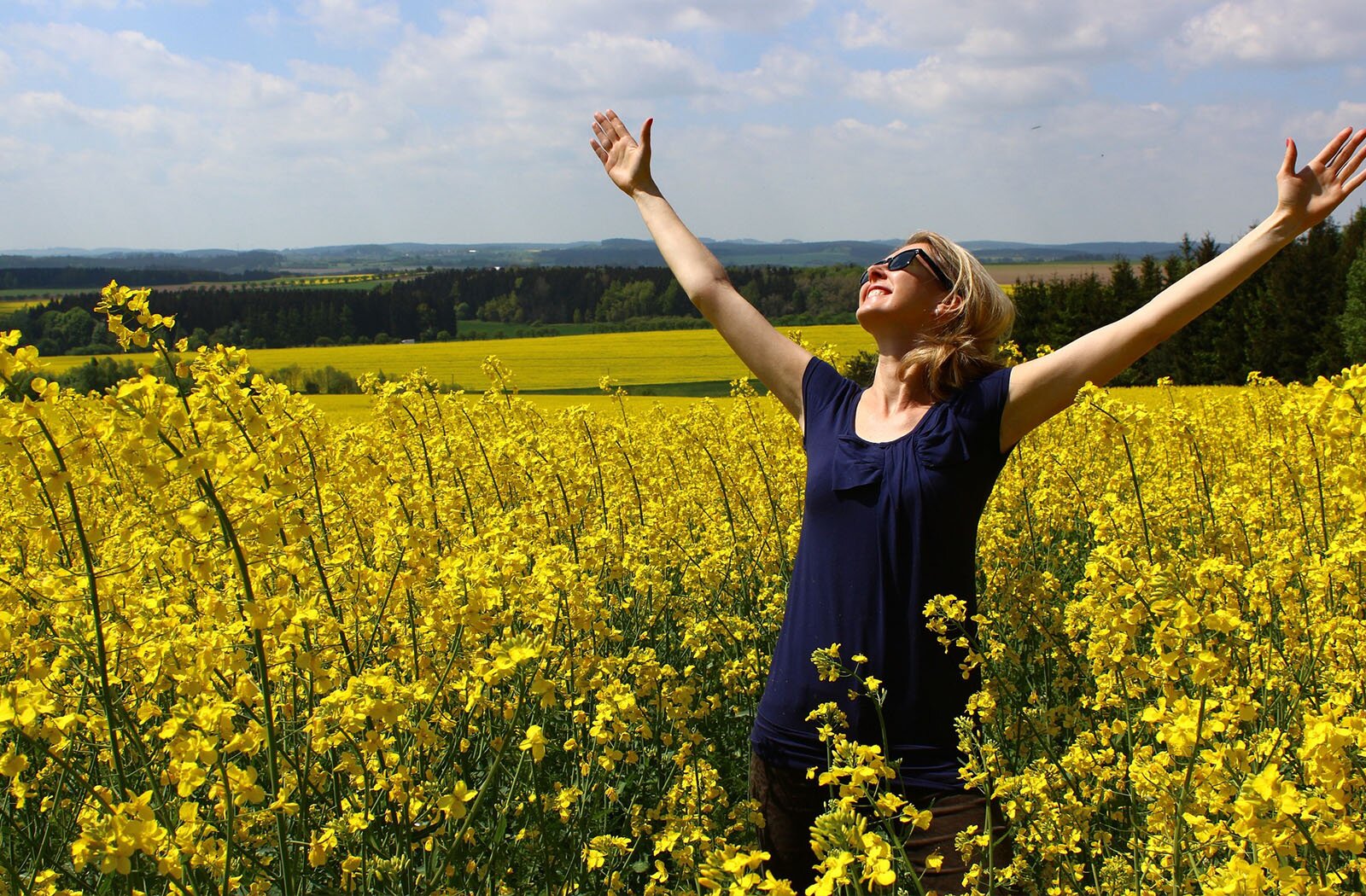 Zdjęcie przedstawia szeroko uśmiechniętą kobietę z uniesionymi rękami do góry, w okularach przeciwsłonecznych. Kobieta stoi na polu rzepaku. 