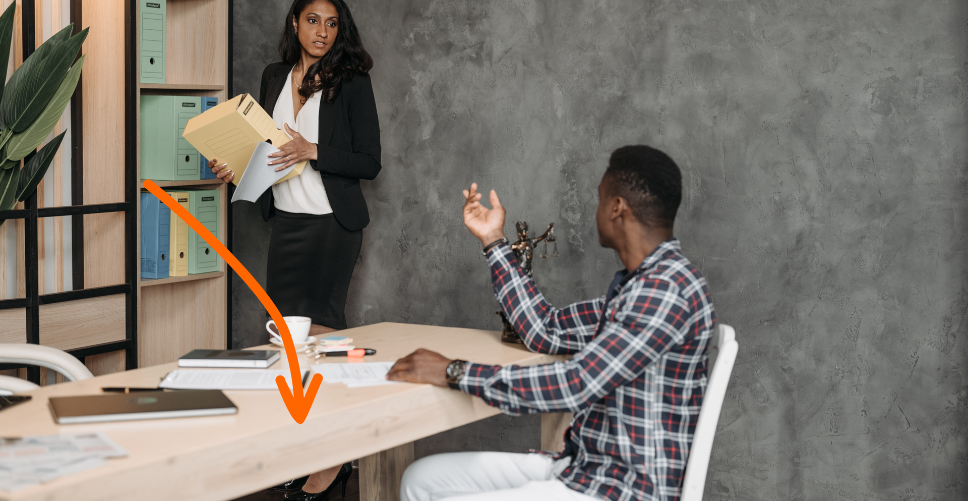 Zdjęcie przedstawia mężczyznę siedzącego za biurkiem oraz kobietę stojącą dalej. Strzałka na ilustracji skierowana jest na biurko.