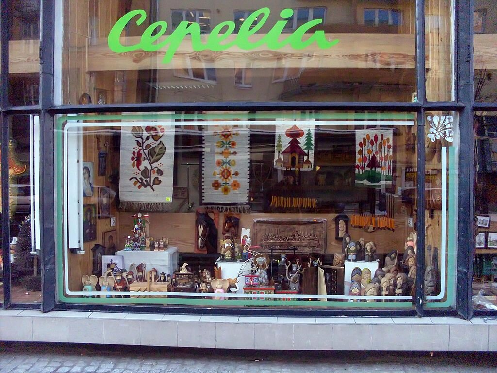 Zdjęcie przedstawia witrynę sklepu Cepelia. Za szybą widoczne są wyroby rękodzielnicze: rzeźby, hafty, obrazy.