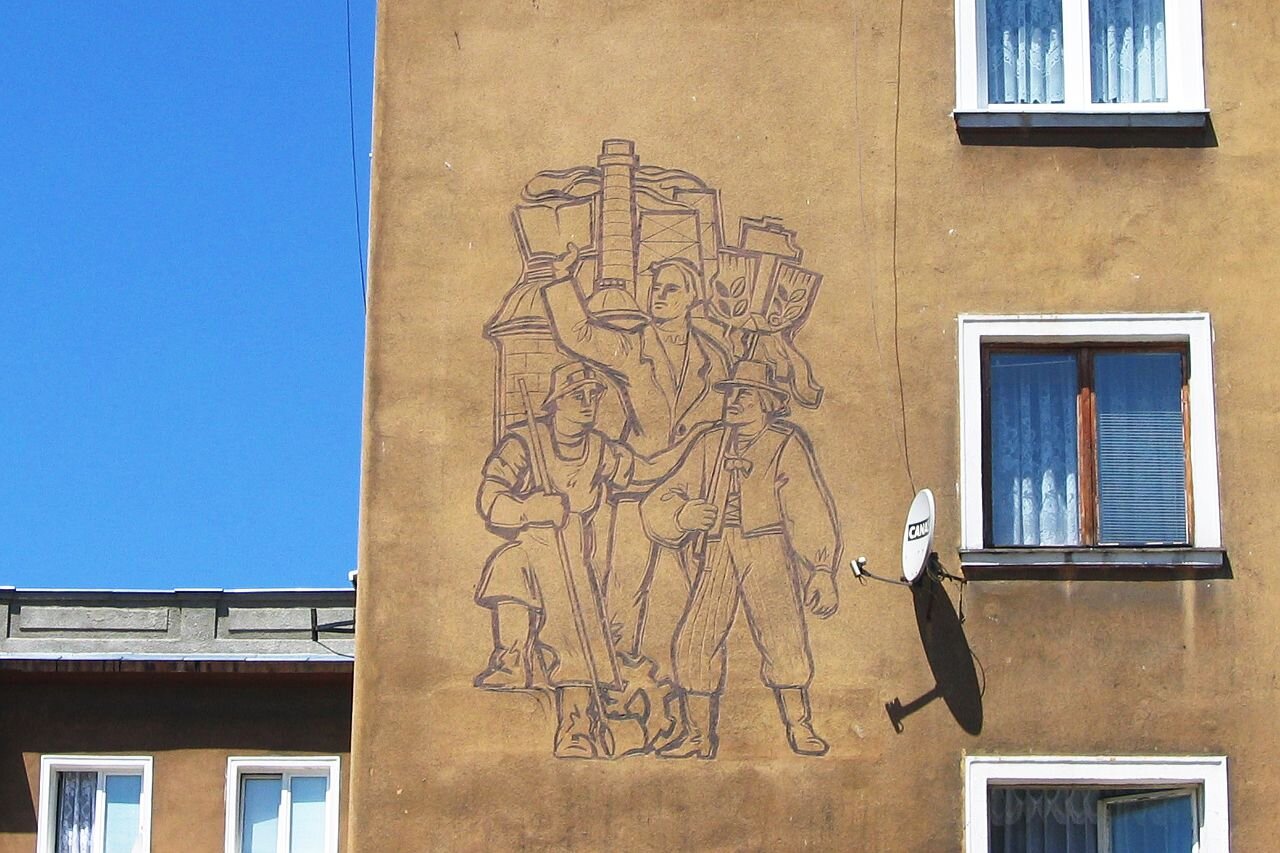 Zdjęcie przedstawia ścianę bloku, na którym znajduje się mural. Przedstawia on trzech mężczyzn. Dwóch z nich jest w strojach robotników, trzeci w stroju chłopskim. W tle, za mężczyznami znajduje się fabryka.