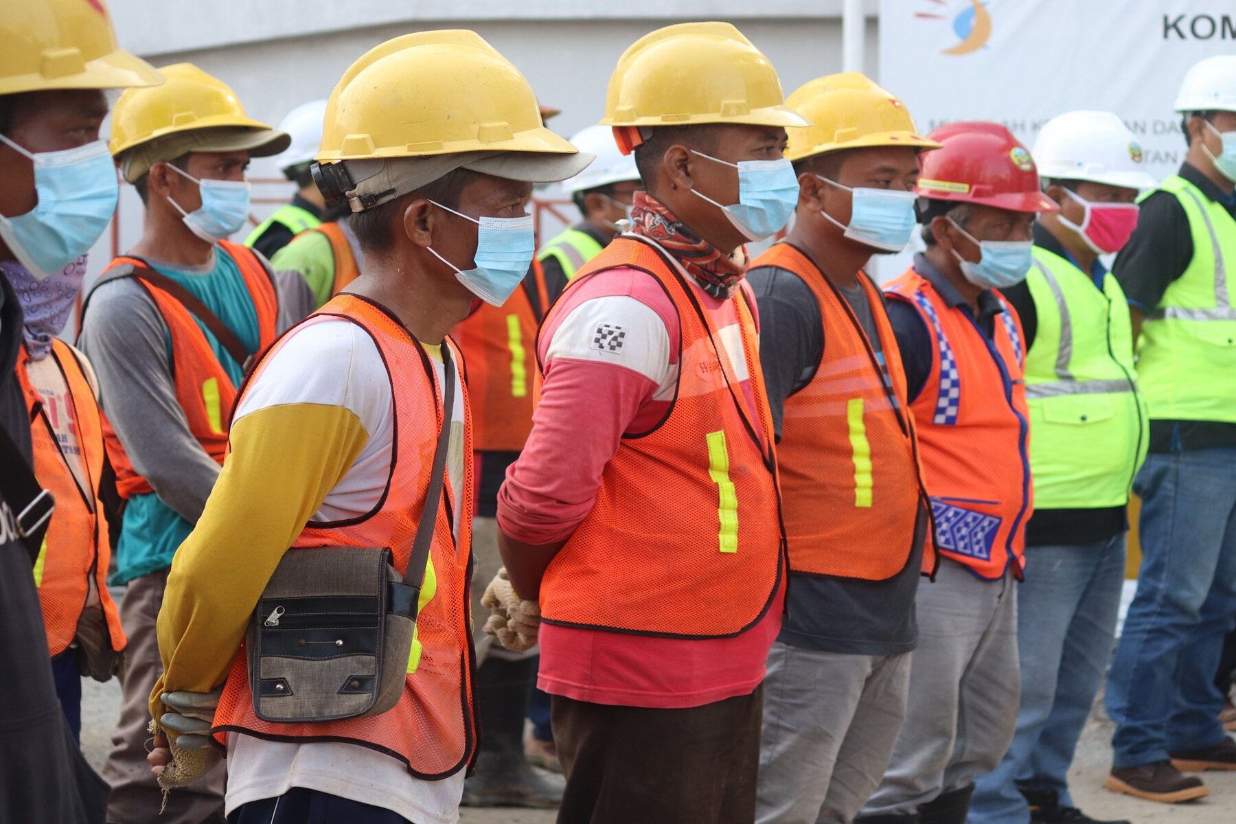 Zdjęcie przedstawia mężczyzn stojących w dwóch rzędach, zwróconych w prawo, w ujęciu z ukosa. Mężczyźni ubrani są w długie spodnie, koszulki z długim rękawem, na które mają założone kamizelki ostrzegawcze w kolorze pomarańczowym lub jaskrawozielonym. Na głowę mają założone kaski ochronne w kolorach: żółtym, czerwonym i białym. Każda osoba ma na twarzy założoną maseczkę ochronną. 