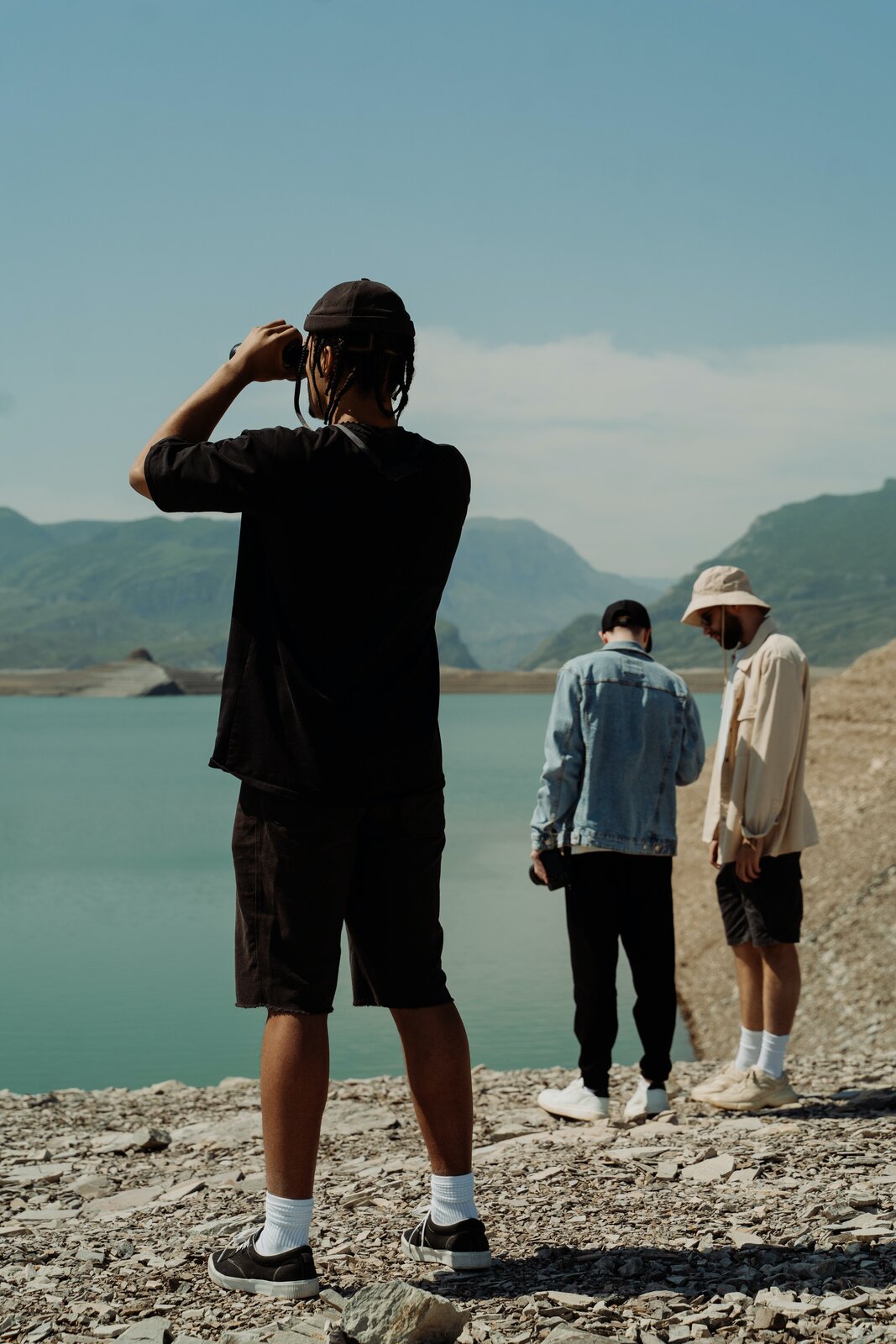 Fotografia przedstawiająca trzech mężczyzn stojących nad brzegiem zbiornika wodnego. Kamienisty brzeg, w oddali wzniesienia.