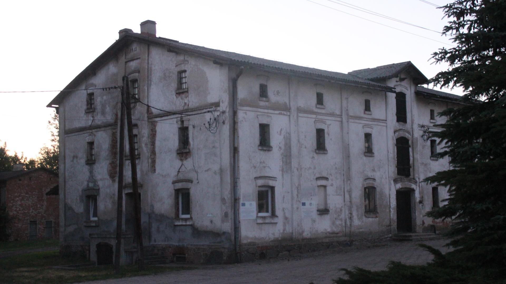Zdjęcie przedstawia dwupiętrowy, biały budynek z lekko skośnym dachem. Okna są małe i rozmieszczone od siebie w równych odległościach. Z lewej strony budynku widoczny jest słup, do którego przywieszony jest kabel. Z prawej strony zdjęcia widoczny jest fragment choinki. Niebo jest jasne, bezchmurne. Zdjęcie zostało wykonane w świetle dziennym.