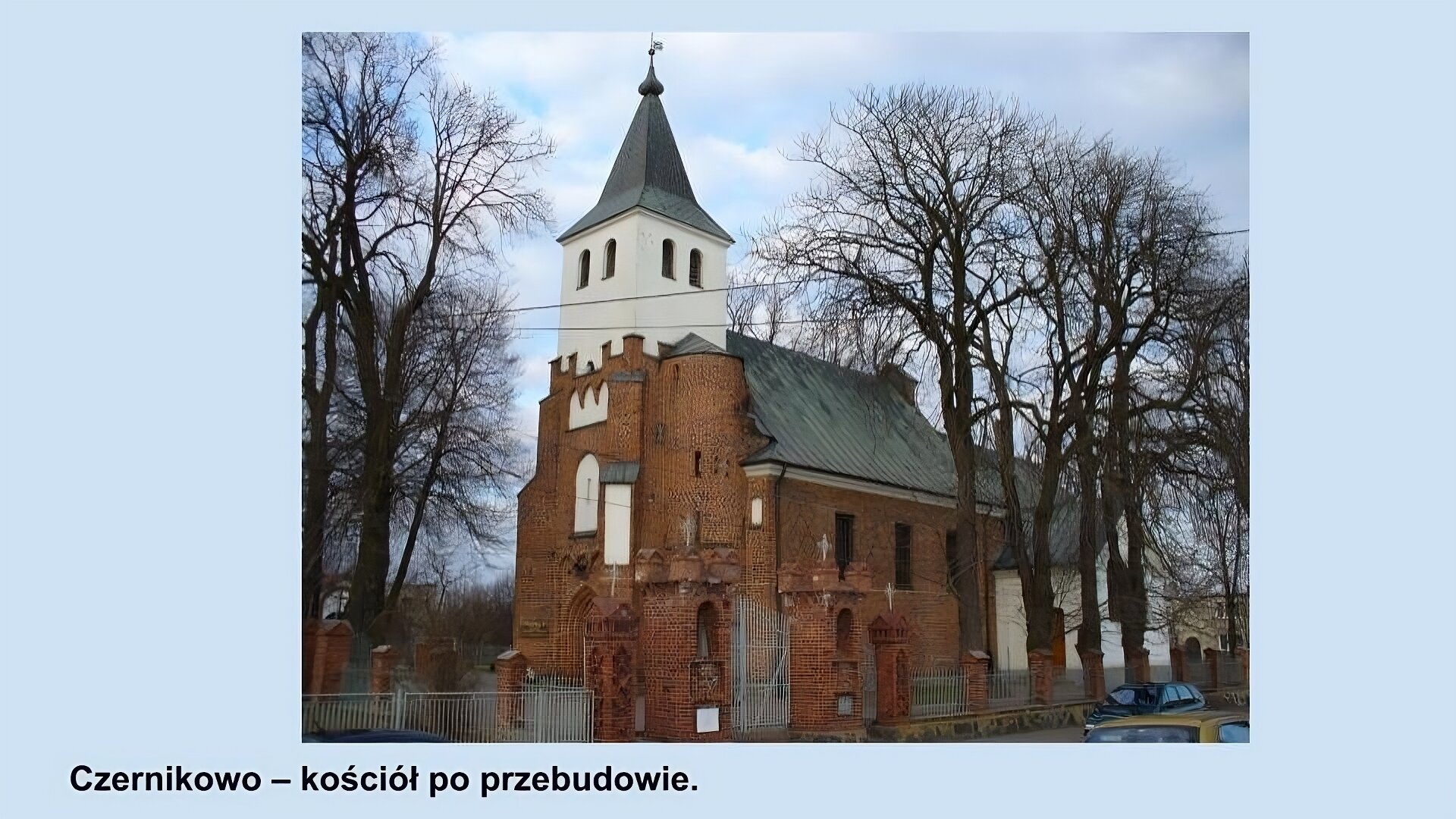 Ilustracja przedstawia kościół w Czernikowie po przebudowie. Ceglany budynek zbudowany na planie prostokąta, ze spadzistym szarym dachem, z białą wieżą, przewyższającą dach, zbudowaną nad głównym wejściem zwieńczoną skośnym, kwadratowym daszkiem. Na około wysokie drzewa bez liści i murowane ogrodzenie z metalowymi, szarymi przęsłami.