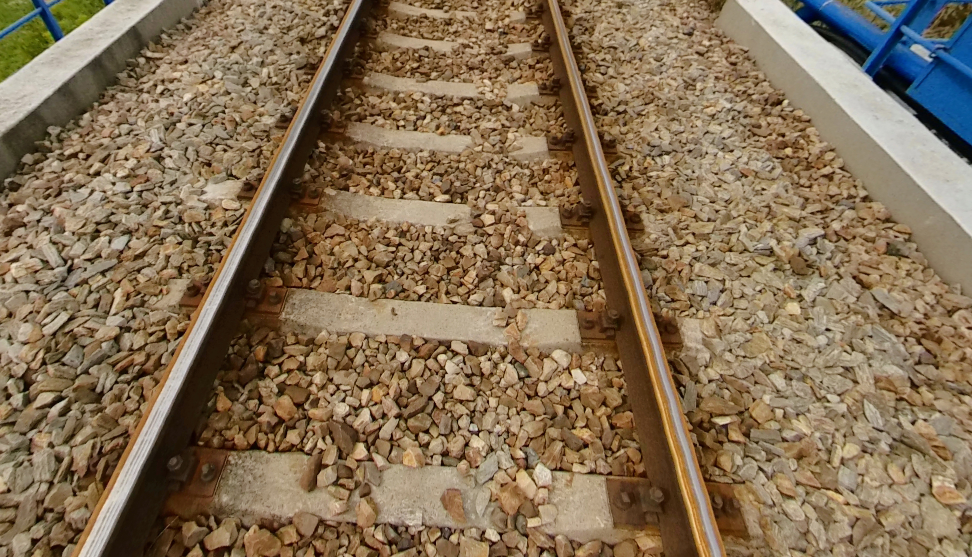 Zdjęcie przedstawia fragment toru kolejowego biegnące po budowli inżynierskiej. Widoczne są szyny na betonowych podkładach. Między podkładami oraz po bokach toru wysypane są kamienie w kolorze jasnobrązowym.