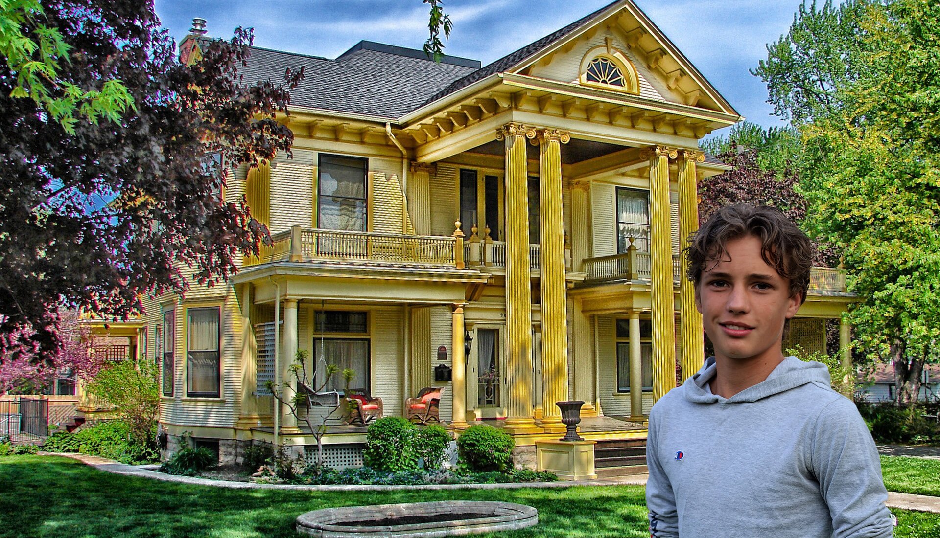 Zdjęcie przedstawia nastoletniego chłopca od pasa w górę. W tle ukazano piętrowy, duży dom, z balkonami oraz filarami ozdobnymi przy wejściu głównym. Dookoła budynku jest wiele zieleni. 