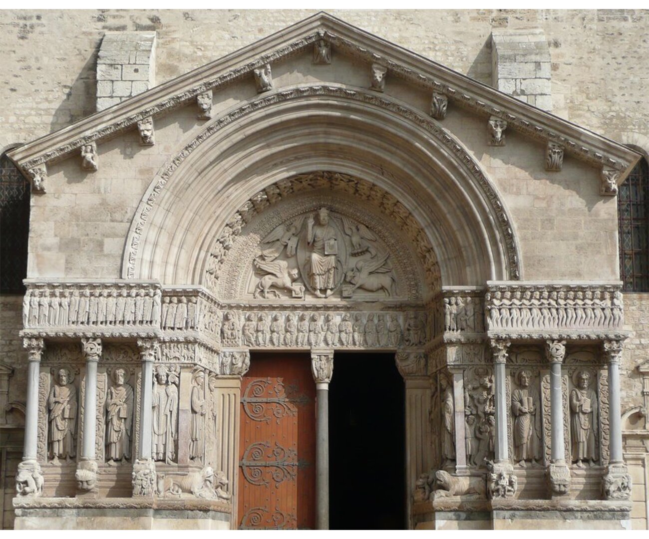 Fotografia barwna przedstawiająca Tympanon kościoła w Arles. Nad bramą główną liczne wyżłobienia, sklepienia.