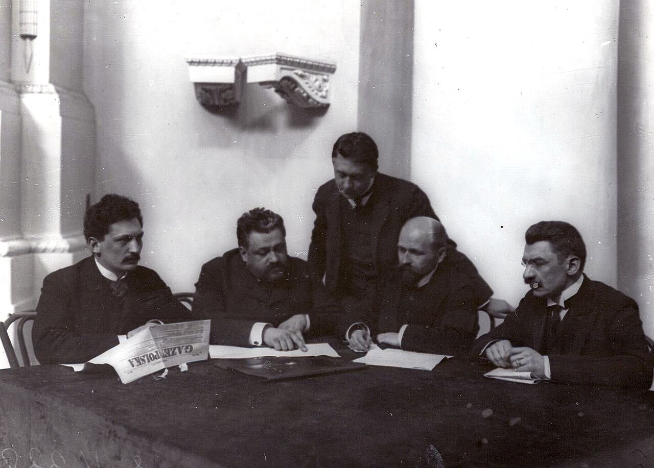 Zdjęcie przedstawia pięciu mężczyzn w garniturach. Siedzą przy stole. Trzymają gazetę i dokumenty.