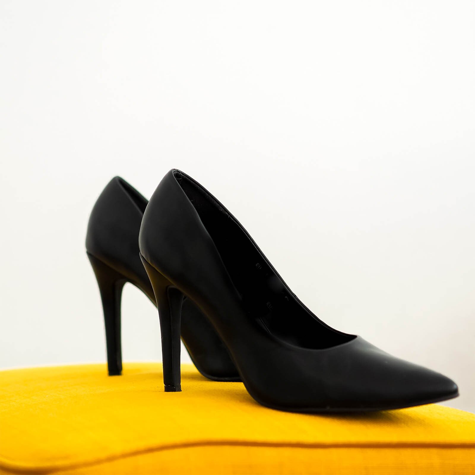 Zdjęcie przedstawia parę czarnego, damskiego obuwia na cienkim obcasie. Buty są ustawione na żółtej poduszce.