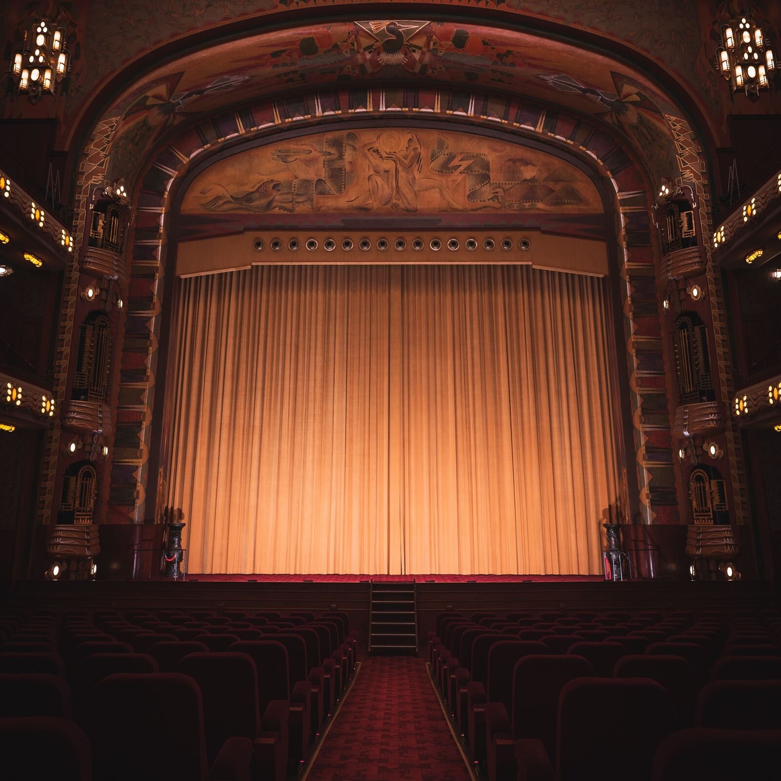 Zdjęcie przedstawia salę teatralną ukazaną z perspektywy widowni, przez środek której jest przejście. Ujęcie przedstawia scenę z zasłoniętą kurtyną, na którą świecą reflektory. W pozostałej części sali panuje półmrok.