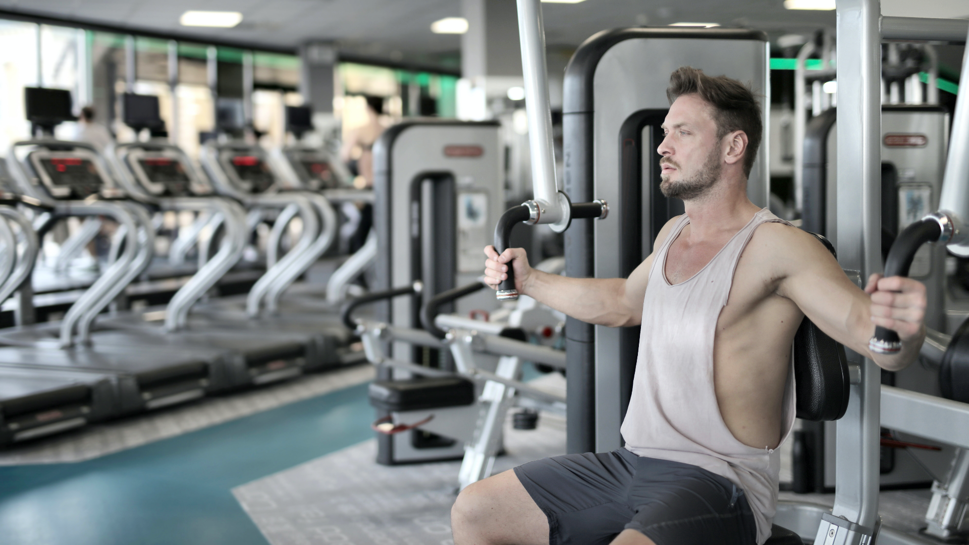 Zdjęcie przedstawia młodego mężczyznę z brodą, który ćwiczy na siłowni. Mężczyzna ubrany jest w koszulkę bez rękawów i krótkie spodenki. Siedzi na urządzeniu do ćwiczeń i rękami trzyma uchwyty. W tle widoczne są ustawione w rzędzie bieżnie.