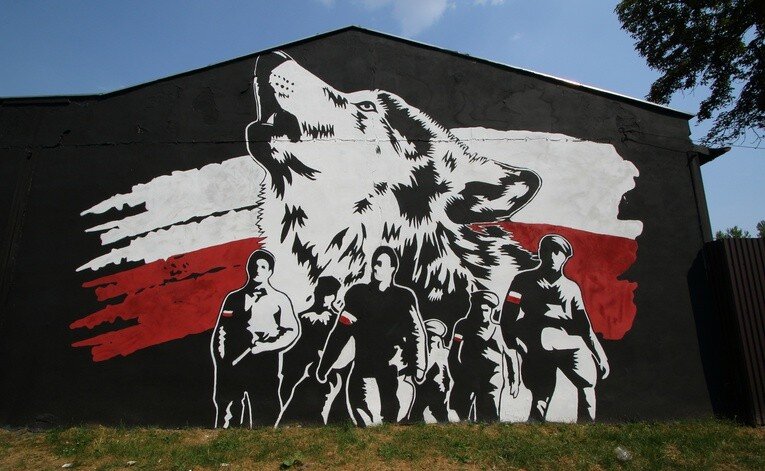 Zdjęcie przedstawia mural na elewacji budynku. Na tle głowy wyjącego wilka narysowani są ludzie z biało‑czerwonymi przepaskami na ramionach. Tłem do całego muralu są dwa pasy w kolorze białym i czerwonym, które wyglądają jak flaga Polski.