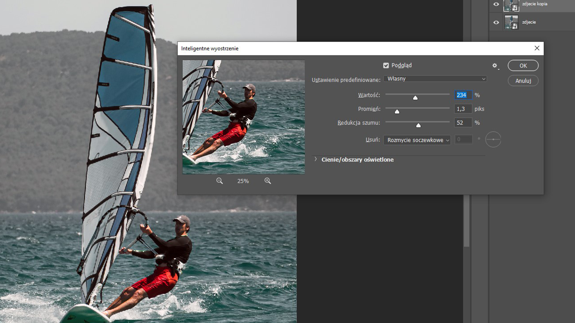 Ilustracja przedstawia okno programu. W obszarze roboczym jest zdjęcie mężczyzny na windsurfingu. Po prawej stronie okno dialogowe o nazwie: Inteligentne wyostrzenie. Zaznaczono: Podgląd, Ustawienia predefiniowane: Własne, Wartość 234%, Promień 1,3 piksela, Redukcja szumu: 52%, Usuń: Rozmycie soczewkowe. Wybrano przycisk: OK. 