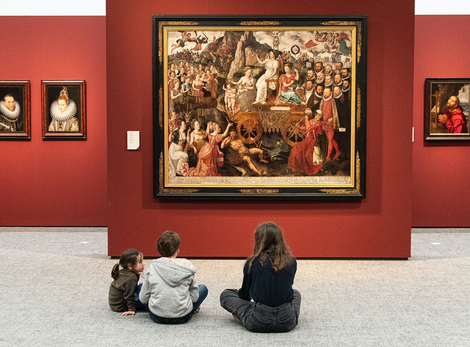 Zdjęcie przedstawia wnętrze galerii sztuki. Widoczne są ściany w kolorze burgundu i wiszące na nich obrazy w ozdobnych ramach. Przed jednym z obrazów przedstawiającym scenkę rodzajową widoczne jest troje dzieci siedzących na podłodze i podziwiających obraz. W tle na ścianie po lewej stronie wiszą portrety, z prawej widoczny mężczyzna składający ręce do modlitwy. Klęczy przed krzyżem. 