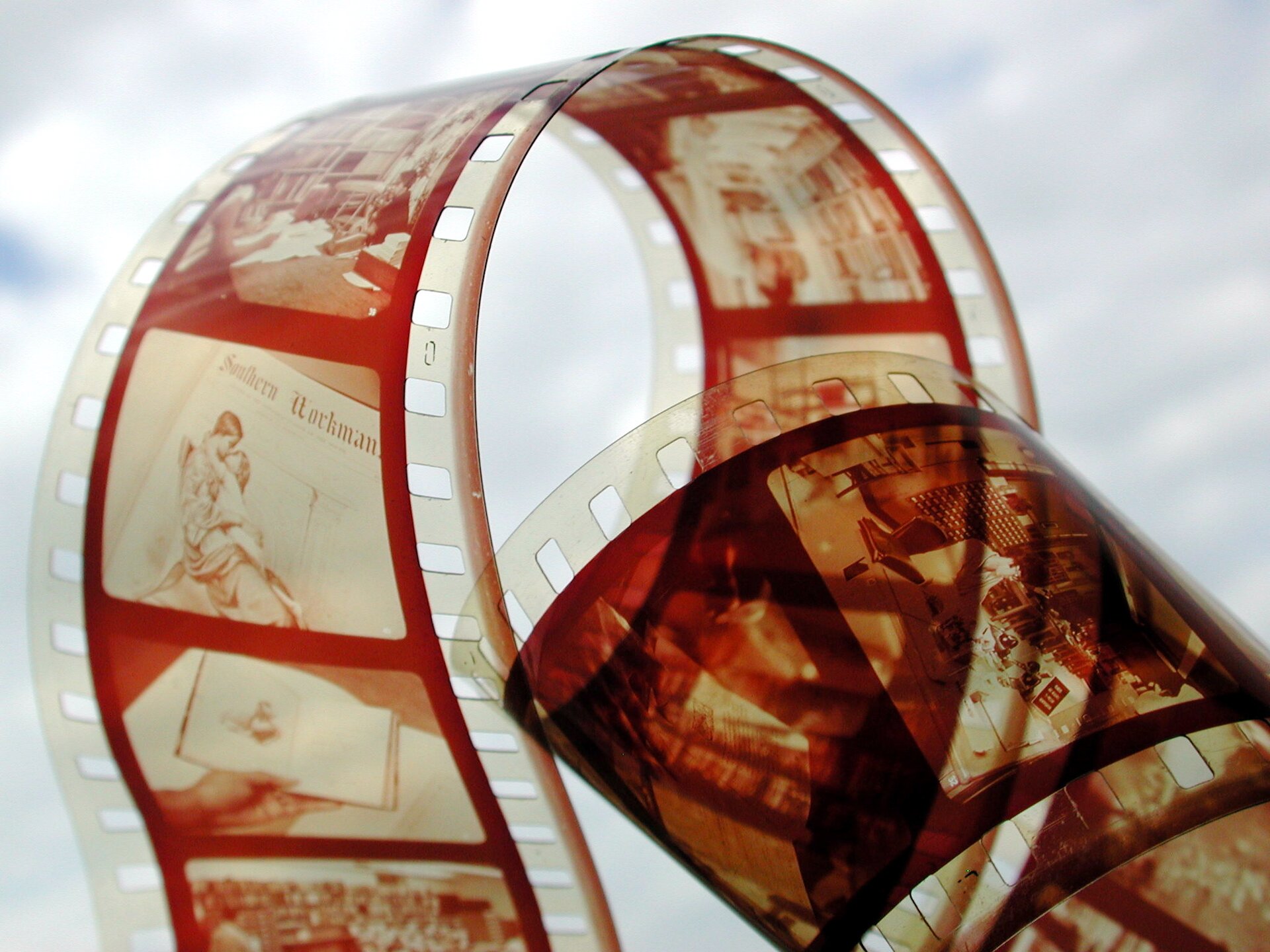 Kliknij, aby powiększyć Na kolorowej fotografii widać zbliżenie na powywijaną czerwono białą taśmę filmową. Tło zdjęcia jest białe i rozmyte.