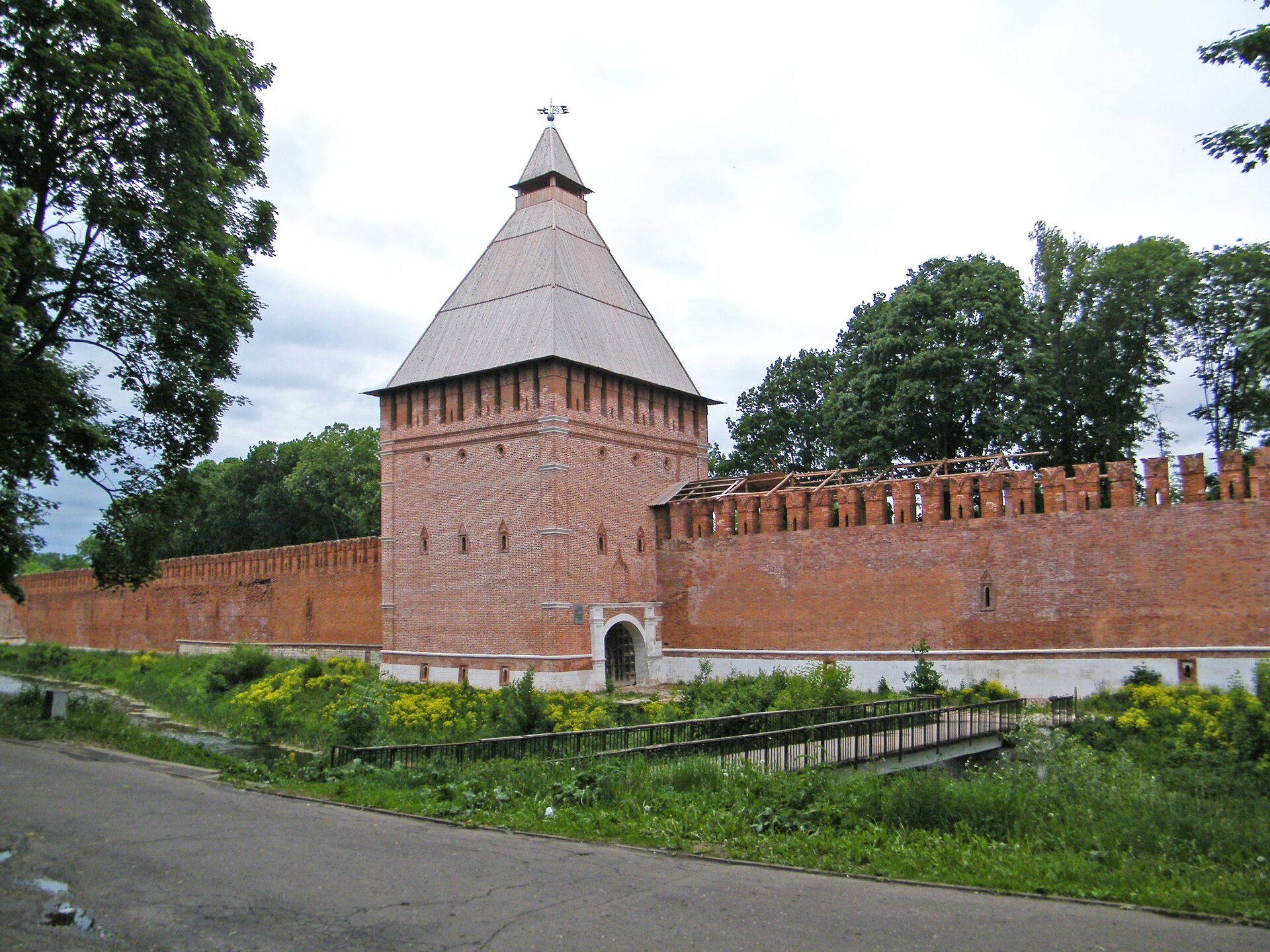 Kliknij, aby powiększyć Fragment murów obronnych twierdzy w Smoleńsku. Widoczna jest też baszta. Mury są zbudowane z czerwonej cegły. Przed murami płynie rzeczka, a przez nią przechodzi most, który prowadzi do drogi.