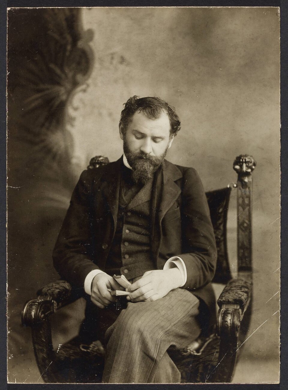 Zdjęcie przedstawia portret mężczyzny w średnim wieku. Mężczyzna siedzi na fotelu, ma opuszczoną głowę i ma spuszczone oczy. W dłoniach trzyma małą książkę. Mężczyzna ma niestarannie ułożone włosy. Nosi brodę. Ubrany jest w garnitur.