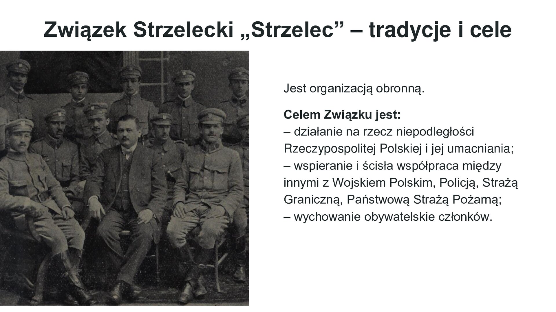 Biały slajd. Z lewej strony czarno‑białe zdjęcie przedstawiające siedzących w dwóch rzędach mężczyzn w mundurach wojskowych. Za nimi, w rzędzie, stoją kolejni żołnierze. U góry slajdu nagłówek: „Związek Strzelecki "Strzelec" - tradycje i cele”. Poniżej, z prawej strony, tekst: „Jest organizacją obronną. Celem Związku jest: działanie na rzecz niepodległości Rzeczypospolitej Polskiej i jej umacniania, wspieranie i ścisła współpraca między innymi z Wojskiem Polskim, Policją, Strażą Graniczną, Państwową Strażą Pożarną; wychowanie obywatelskie członków”.