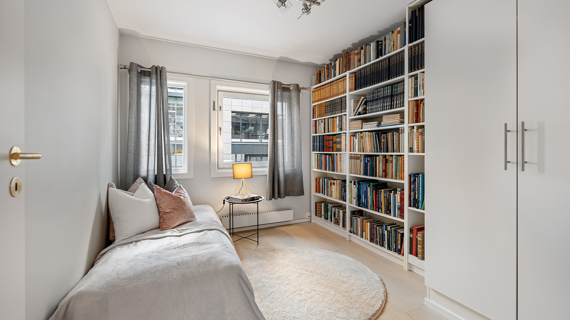 Zdjęcie przedstawia małą sypialnią, w której nie ma zbyt wielu mebli. Na ścianie naprzeciwko obserwatora znajduje się okno z zasłonami, ale bez firan. Na ścianie po lewej stronie okna znajduje się pojedyncze łóżko z dwiema małymi poduszkami. Po prawej stronie okna stoi duży regał wypełniony książkami oraz dwudrzwiowa  szafa. Pod oknem stoi mały stolik, a na nim lampa.