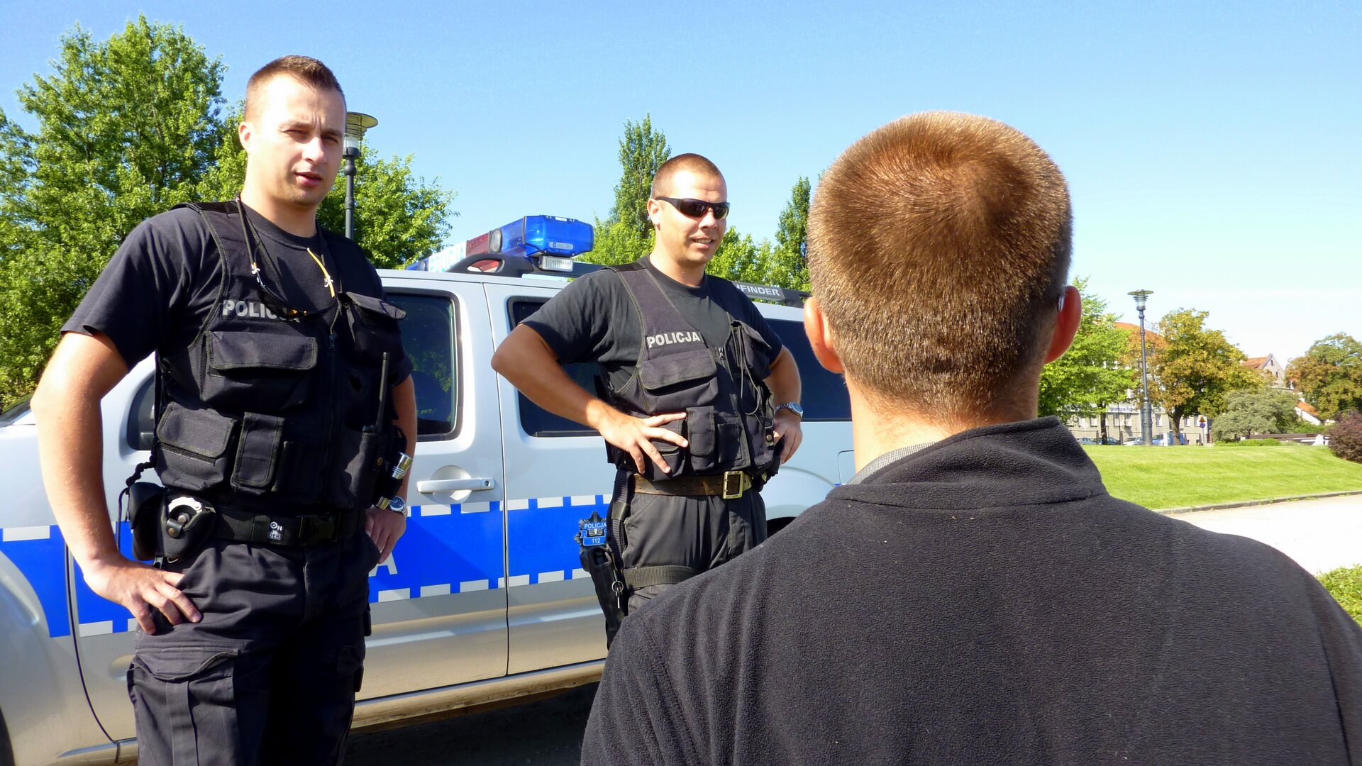 Zdjęcie, które przedstawia patrol policji. Dwóch policjantów, którzy rozmawiają z mężczyzną. Za policjantami zaparkowane auto policyjne. Policjanci ubrani są w mundury służbowe, koszula polo z krótkim rękawem, spodnie służbowe, kamizelka ostrzegawcza z napisem policja, pas policyjny do przenoszenia broni krótkiej, przy nodze pałka służbowa. 
