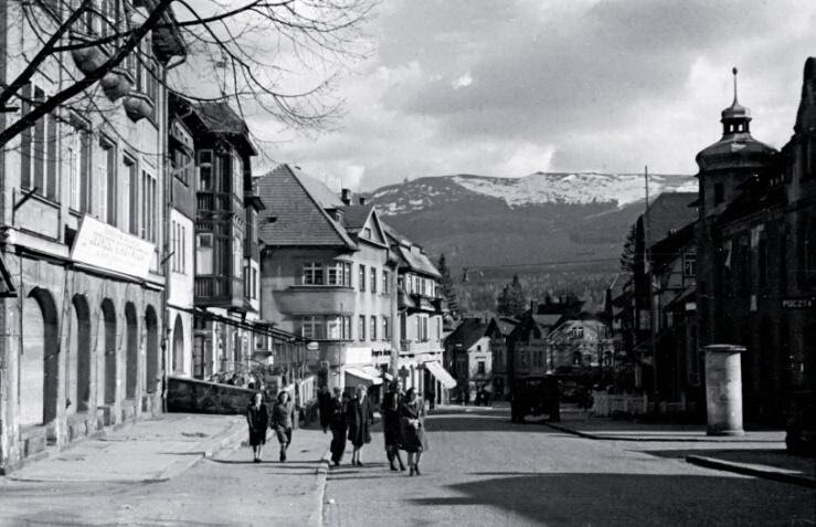 Zdjęcie przedstawia ulicę w Szklarskiej Porębie, przy której stoją liczne kamienice. Ulicą idzie grupa ludzi. Na drugim planie widać Karkonosze i Szrenicę.