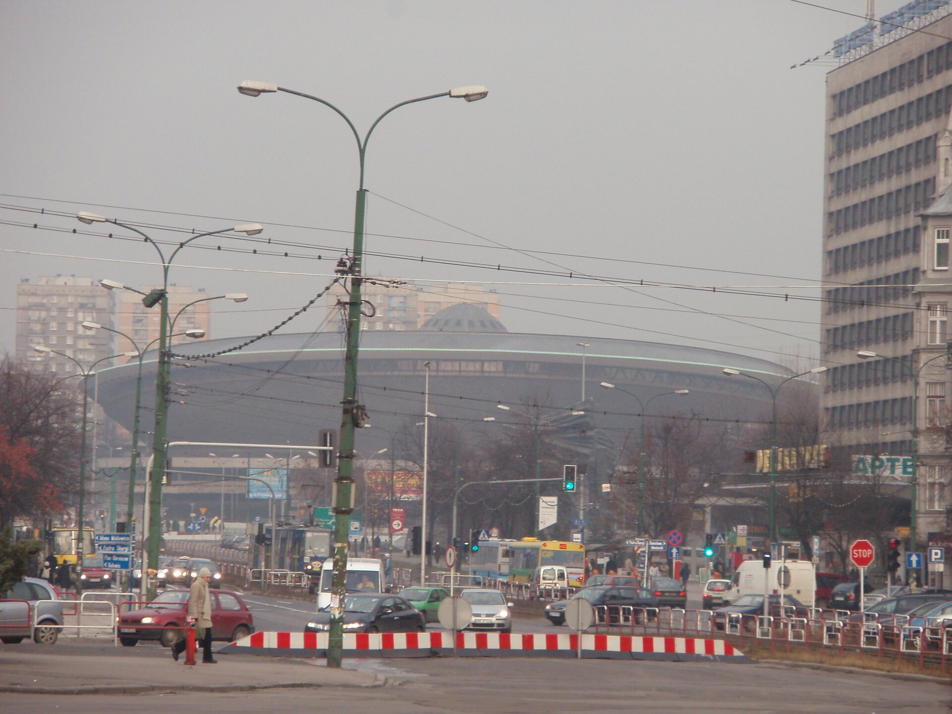 Fotografia katowickiego Spodka wykonana na ulicy w odległości kilkudziesięciu metrów przed nim. Krajobraz jest wypełniony smogiem przypominającym szarą mgłę. Ograniczona widoczność.