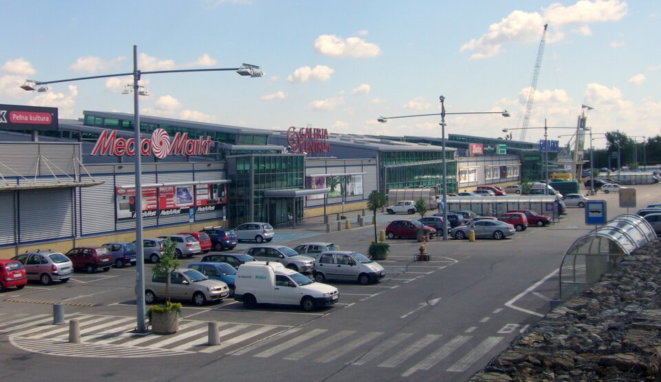 Na zdjęciu znajduje się rozległa galeria handlowa z dużym parkingiem. Galeria Centrum rozciąga się od prawej strony kadru przez całe zdjęcie. Jest to dość niski budynek z wieloma reklamami na elewacji i nad budynkiem. Widoczne są reklamy sklepów Empik, Media Markt i Bata. Na fasadzie szare płyty przypominające zasłonięte srebrne żaluzje. Cały budynek utrzymany jest w kolorystyce szarej, ciemnoszarej i zielonkawej. Okna są ciemno‑zielone, a najwięcej jest ich przy wejściu do galerii, nad którym znajduje się zadaszenie. Przed galerią znajduje się duży parking, na którym stoją samochody niezajmujące całej jego powierzchni. Przez parking przebiega oznakowane na ulicy przejście dla pieszych. Na parkingu znajdują się trzy szare wysokie latarnie z lampami przywieszonymi do szerokich ramion. Na dwóch miejscach widoczne jest zadaszone pleksą miejsce na wózki sklepowe. Po prawej stronie kadru fragment przystanku autobusowego ze znakiem drogowym. W tle niebieskie niebo z pojedynczymi chmurami.