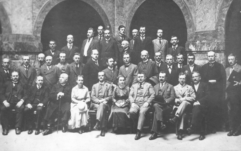 Zdjęcie przedstawia grupę kilkudziesięciu osób pozującą do fotografii, ustawionych w trzy rzędy. W grupie dominują mężczyźni, tylko w pierwszym rzędzie widoczne są dwie kobiety.  Mężczyźni ubrani są w garnitury, a kobiety w długie suknie.