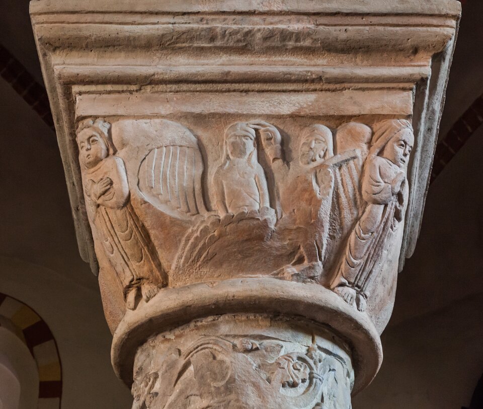 Ilustracja przedstawia fotografię głowicy z kolumny cnót, znajdującej się w kościele św. Trójcy w Strzelnie. Kostkowa głowica ukazuje grupy aniołów, stojących w pozach modlitewnych ze złożonymi lub skrzyżowanymi rękoma. Niektóre z aniołów grają na instrumentach. Od góry głowica zwieńczona jest uskokowo. 