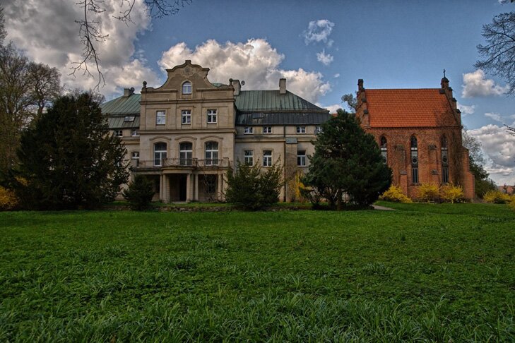 Na zdjęciu kilkupiętrowy budynek. Jest to pałac. Do wejścia prowadzą kolumny. Ma wysoki dach. Na środku znajduje się wystająca część budynku z dekoracyjnym zwieńczeniem. Po prawej stronie pałacu znajduje się kościół. Przed pałacem jest trawa.
