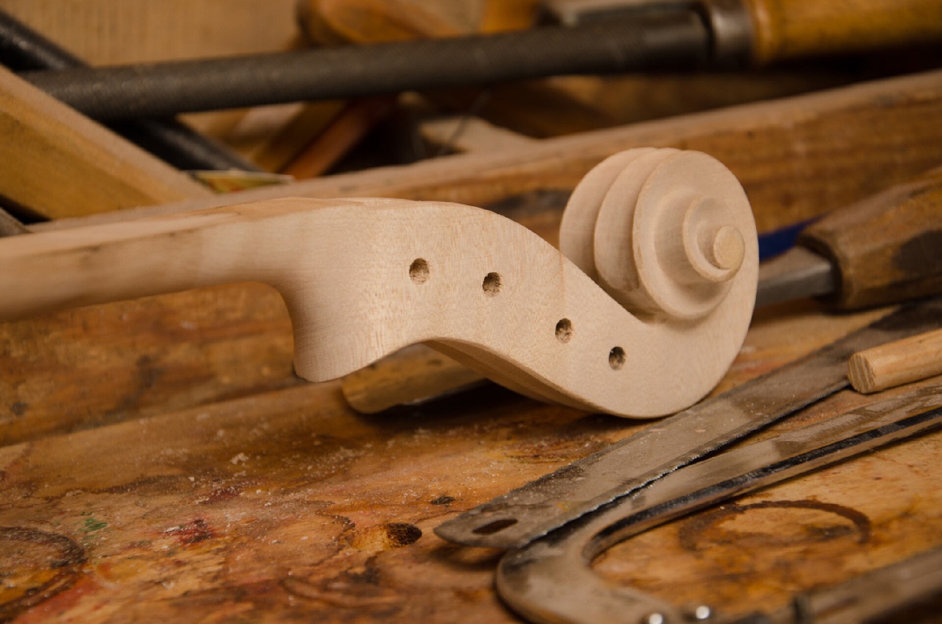 Na zdjęciu szyjka i główka skrzypiec w trakcie pracy lutnika. Elementy są niepolakierowane, w główce nie są jeszcze umieszczone kołki. W tle widać narzędzia lutnicze.