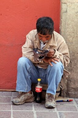 Czarno-białe zdjęcie przedstawia biednego mężczyznę żebrzącego na ulicy. Mężczyzna jest przycupnięty na chodniku, oparty o słup. Ubrany jest w garnitur, a na głowie ma czapkę. 