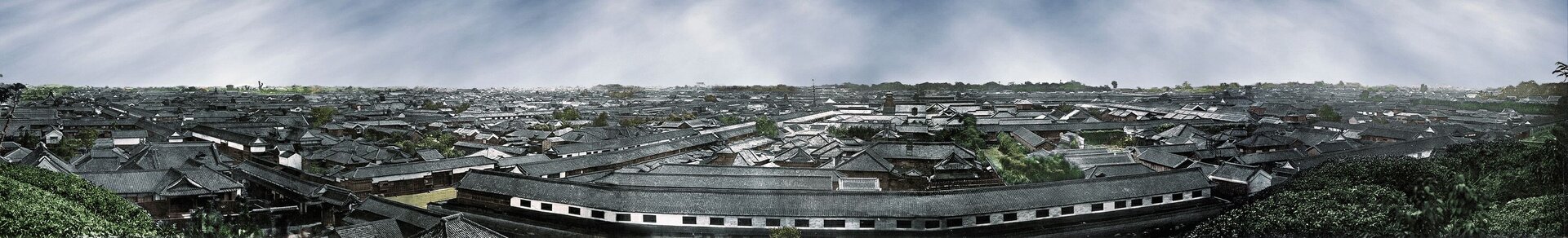 Zdjęcie przedstawia panoramę miasta Tokio w drugiej połowie XIX wieku. Szereg zabudowań z dwuspadowymi, szarymi dachami, wybudowanych w nieregularnym układzie. Na pierwszym planie długi budynek z białą ścianą boczną i szeregiem otworów okiennym w kształcie prostokąta. Po obu krawędziach fotografii widoczne zieleńce wzniesienia, z którego wykonywano fotografię. 