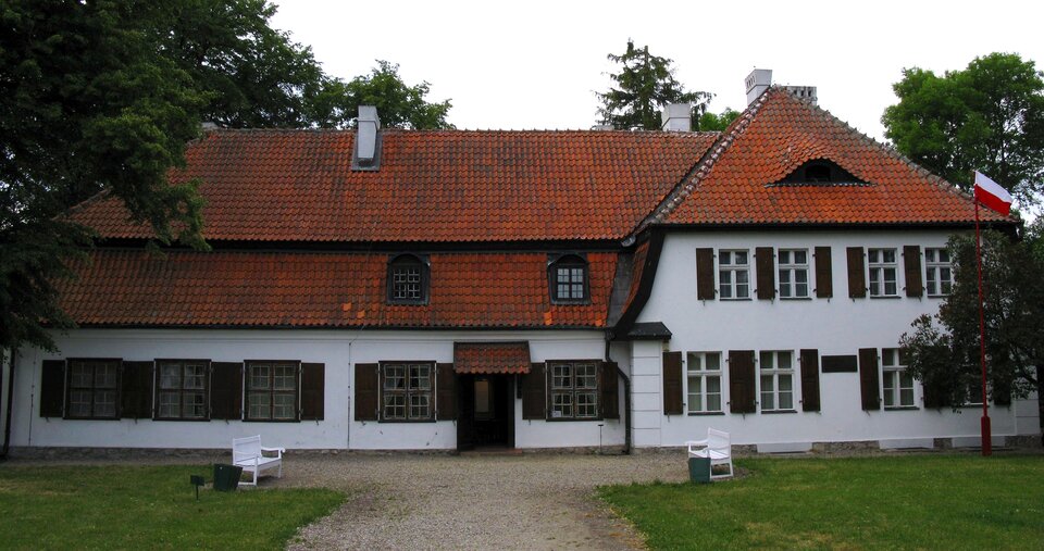 Zdjęcie przedstawia biały, piętrowy budynek z pomarańczowym, skośnym dachem. Z prawej strony budynku znajduje się alkierz, na ścianie którego znajdują się dwa piętra okien z drewnianymi okiennicami. Na dachu znajduje się lukarna z małym oknem. Z lewej strony budynku widoczne są okna na parterze z drewnianymi okiennicami oraz drewniane drzwi, z daszkiem umieszczonym nad nimi. Z obu stron budynku widoczne są zielone, duże drzewa, a z prawej dodatkowo powiewa biało‑czerwona flaga. Przed budynkiem rośnie zielona trawa, a do wejścia prowadzi piaskowa droga. Po obu jej stronach umieszczone zostały białe ławki. Niebo jest jasne. Zdjęcie zostało wykonane w świetle dziennym.