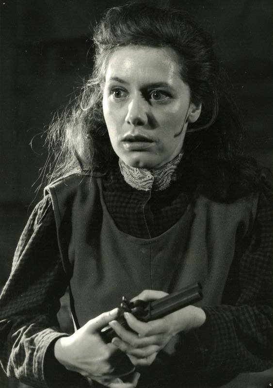 Czarno‑biała fotografia prezentuje młodą kobietę w ujęciu do pasa. Kobieta ma długie włosy, ubrana jest w suknię. W dłoniach trzyma pistolet. Na jej twarzy maluje się przerażenie.