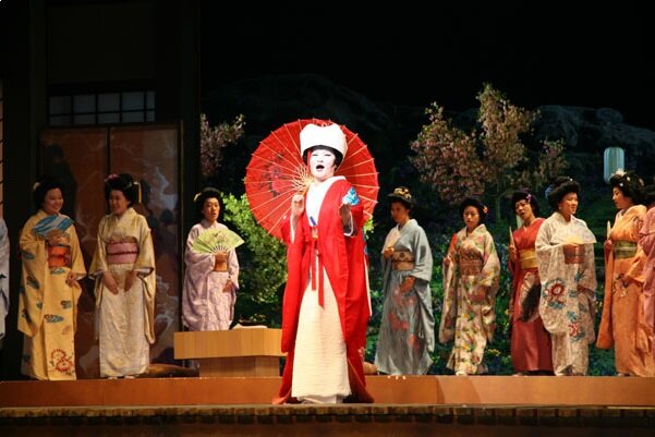 Scena z opery Madame Butterfly. Na operowej scenie jest dziewięć kobiet w tradycyjnych strojach japońskich. Jedna z nich jest wysunięta na pierwszy plan; ma czerwona szatę i białe nakrycie głowy. W prawej dłoni trzyma czerwoną parasolkę z ozdobnym japońskim wzorem. Pozostałe kobiety stoją na drugim planie, mają różnokolorowe kimona i są bez nakrycia głowy.