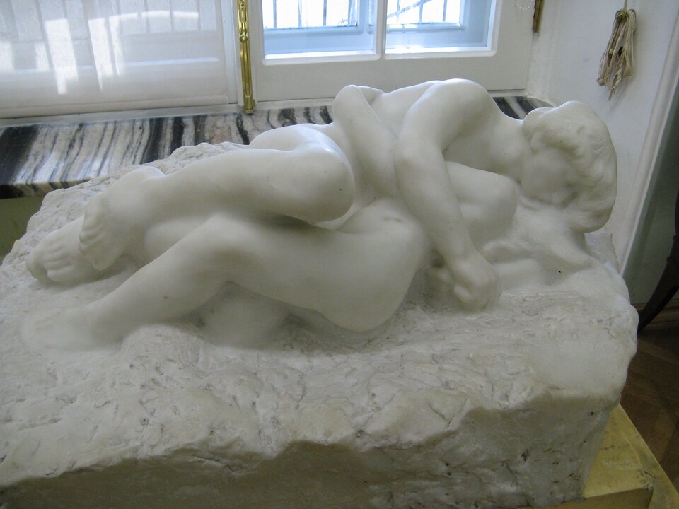 Zdjęcie przedstawia marmurową rzeźbę wyobrażającą obejmującą się parę kochanków. Kobieta i mężczyzna są nadzy. Obejmują się ramionami. Ich nogi są ugięte i splecione. Twarz kobiety jest zasłonięta. Mężczyzna całuje jej szyję, zamykając oczy.