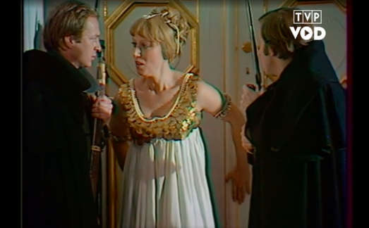 Zdjęcie przedstawia trzy postaci. Po lewej stronie stoi mężczyzna w sile wieku. Postać ukazuje prawy profil. Mężczyzna ma jasne, krótkie włosy. Jego czoło jest wysokie i gładkie, brwi jasne, oczy zwężone, nos niewielki, a usta małe. Ma gładko ogolone policzki i podbródek. W lewej dłoni zaciska strzelbę. Ubrany jest w ciemny płaszcz z postawionym kołnierzem. Pośrodku stoi młoda kobieta. Postać ukazuje lewy półprofil owalnej twarzy. Jej czoło jest przykryte grzywką. Kobieta ma jasne brwi, zwężone oczy, prosty nos i lekko otwarte usta. Jej długie włosy są upięte z tyłu głowy. Fryzura jest ozdobiona wąskimi wstążkami. Patrzy na mężczyznę po lewej. Rękoma przymyka drzwi stojące za jej plecami. Jest ubrana w luźną koszulę bez rękawów. Po prawej stronie stoi mężczyzna w sile wieku. Postać ukazuje lewy ćwierćprofil. Włosy mężczyzny są krótkie i ciemne. Ma bokobrody. W dłoniach trzyma bagnet. Jest ubrany w ciemny płaszcz. Drzwi stojące za ich plecami są bogato zdobione złotymi detalami.