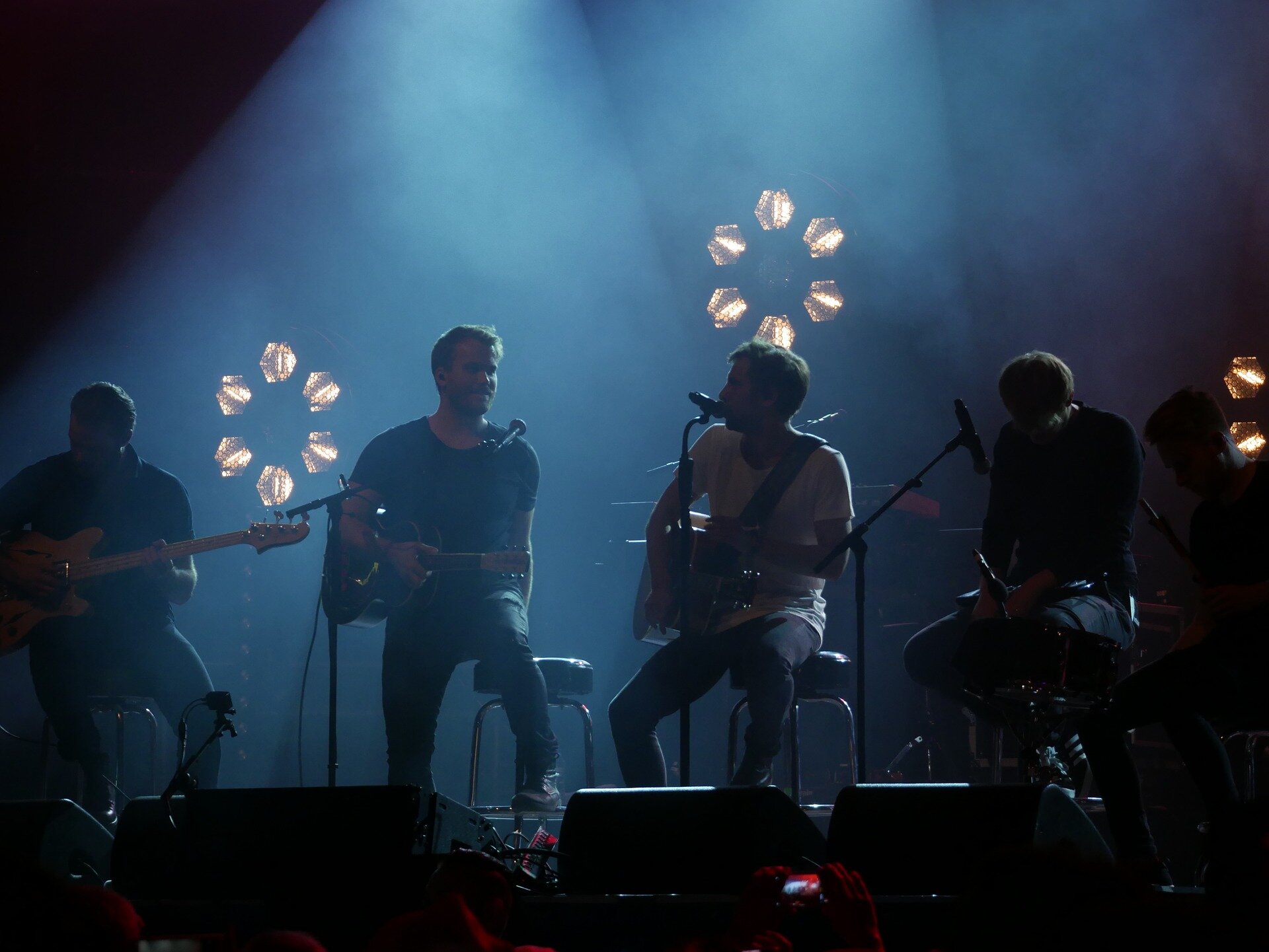 Zdjęcie przedstawia pięcioosobowy zespół muzyczny na scenie podczas koncertu. Składa się on z gitarzystów, perkusisty oraz wokalisty.