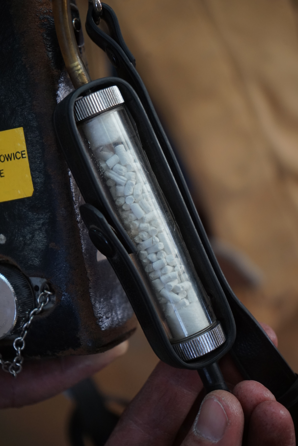 Zdjęcie przedstawia fragment metanomierz. Centralnym elementem zdjęcia jest walcowaty zbiornik. Zbiornik wypełniony jest białymi granulkami o cylindrycznym kształcie. Jest to środek suszący, zapobiegający dostawaniu się wilgoci do wnętrza komór pomiarowych. Zbiornik znajduje się pomiędzy skórzanym paskiem. Do górnej części zbiornika dochodzi gumowa rurka.