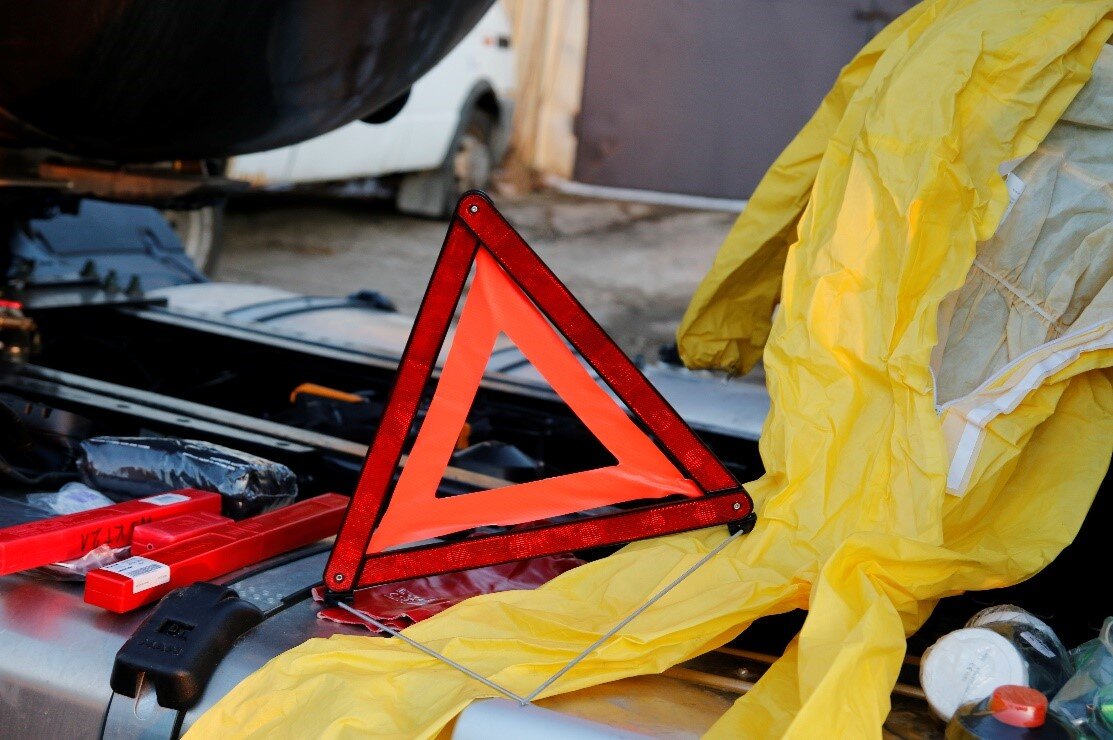 Ilustracja przedstawia składany trójkąt ostrzegawczy. Trójką ostrzegawczy składa się z dwóch trójkątów równobocznych, Pierwszy koloru czerwonego a w nim wpisany mniejszy trójkąt koloru pomarańczowego. W tle widać konieczne wyposażenie samochodowe.