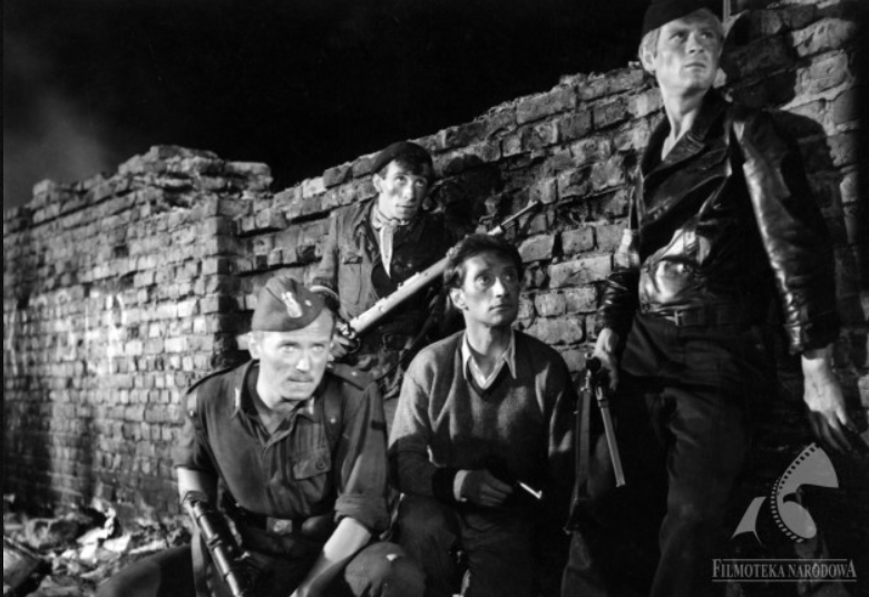 Zdjęcie przedstawia ceglany mur, przy którym znajduje się czterech mężczyzn z karabinami. Jeden z mężczyzn ubrany jest w polski mundur z okresu drugiej wojny światowej. Trzech pozostałych mężczyzn ubranych jest w cywilne ubrania.