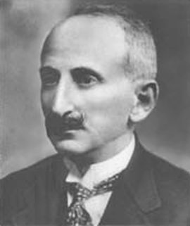 Fotografia przedstawia portret mężczyzny w średnim wieku. Mężczyzna ma krótkie włosy i wąsy. Ubrany jest w białą koszulę, krawat, ciemną marynarkę. 